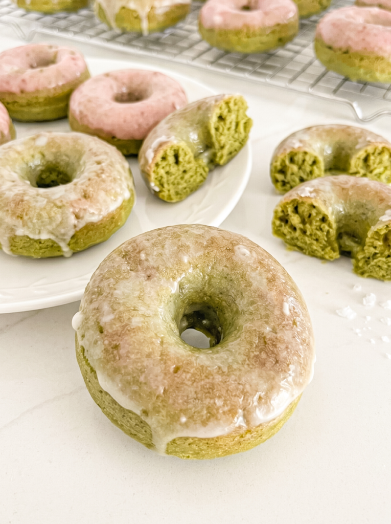Baked matcha donuts, some with white glaze and some with pink glaze, are arranged on a plate and a cooling rack.