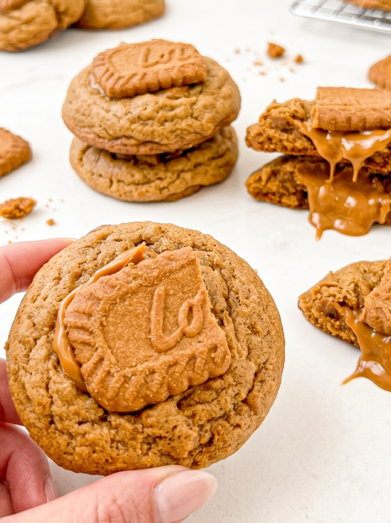 Hand holding a round gingerbread cookie topped with Biscoff spread and a piece of Biscoff cookie, with more cookies in the background.