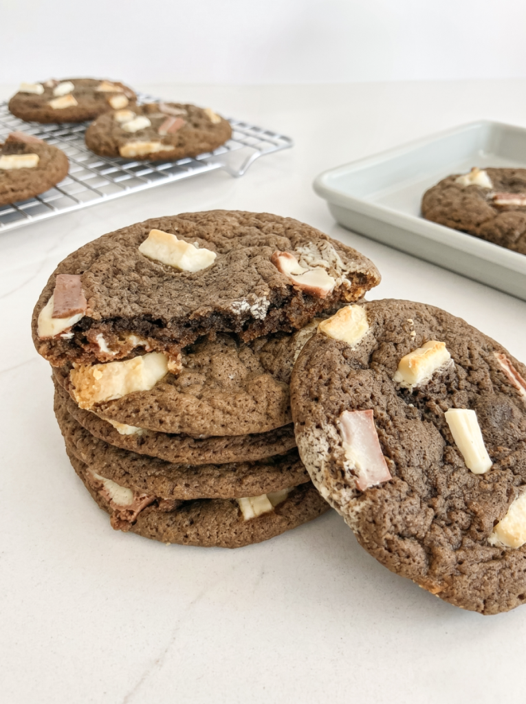 A stack of dark brown cookies with white and pinkish chunks sits on a white surface, with one cookie broken open and more cookies in the background.