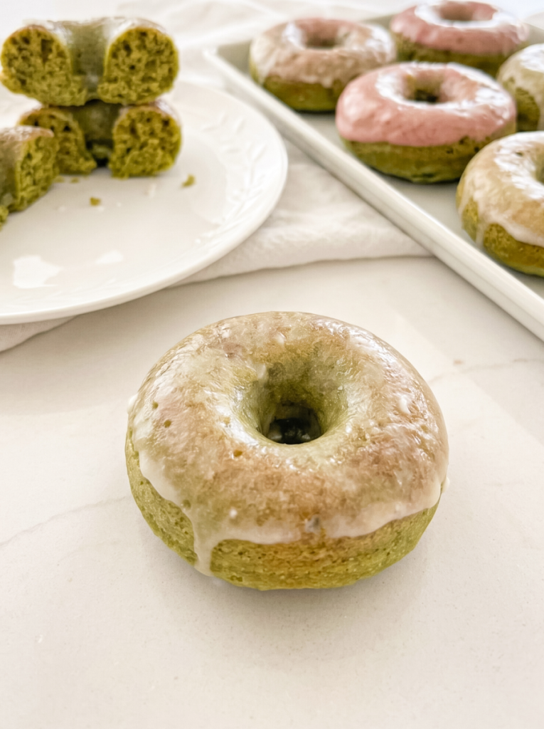 A green matcha donut with white glaze is in the foreground, with stacked donut halves on a plate and more glazed donuts on a tray behind it.