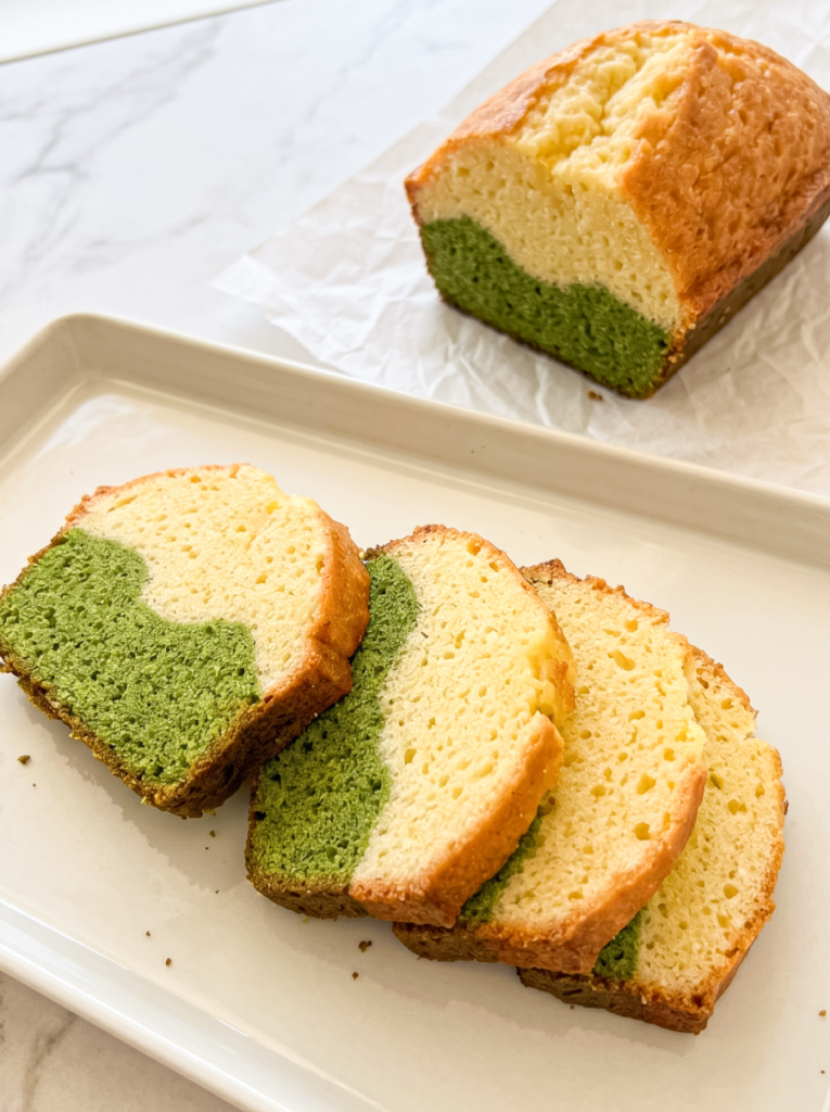 A Matcha Latte Pound Cake loaf is sliced, showing green and pale yellow marbled sections on a white plate and parchment paper.
