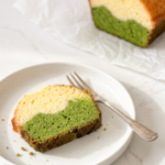 A slice of matcha latte pound cake with distinct green and cream layers is on a white plate with a fork, with the loaf in the background.