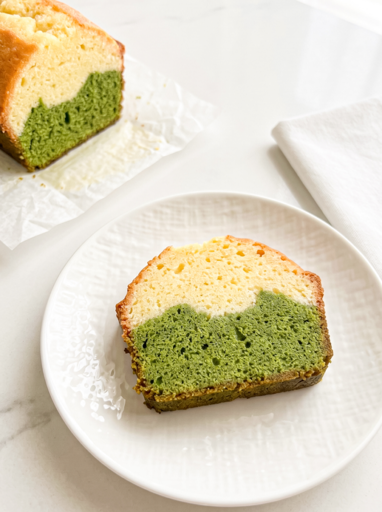 A slice of matcha latte pound cake with green and cream layers is on a white plate, with the loaf in the background.