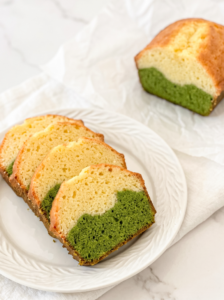 Four slices of a matcha latte pound cake with green bottom and light yellow top layers are arranged on a white plate, with the rest of the loaf in the background.