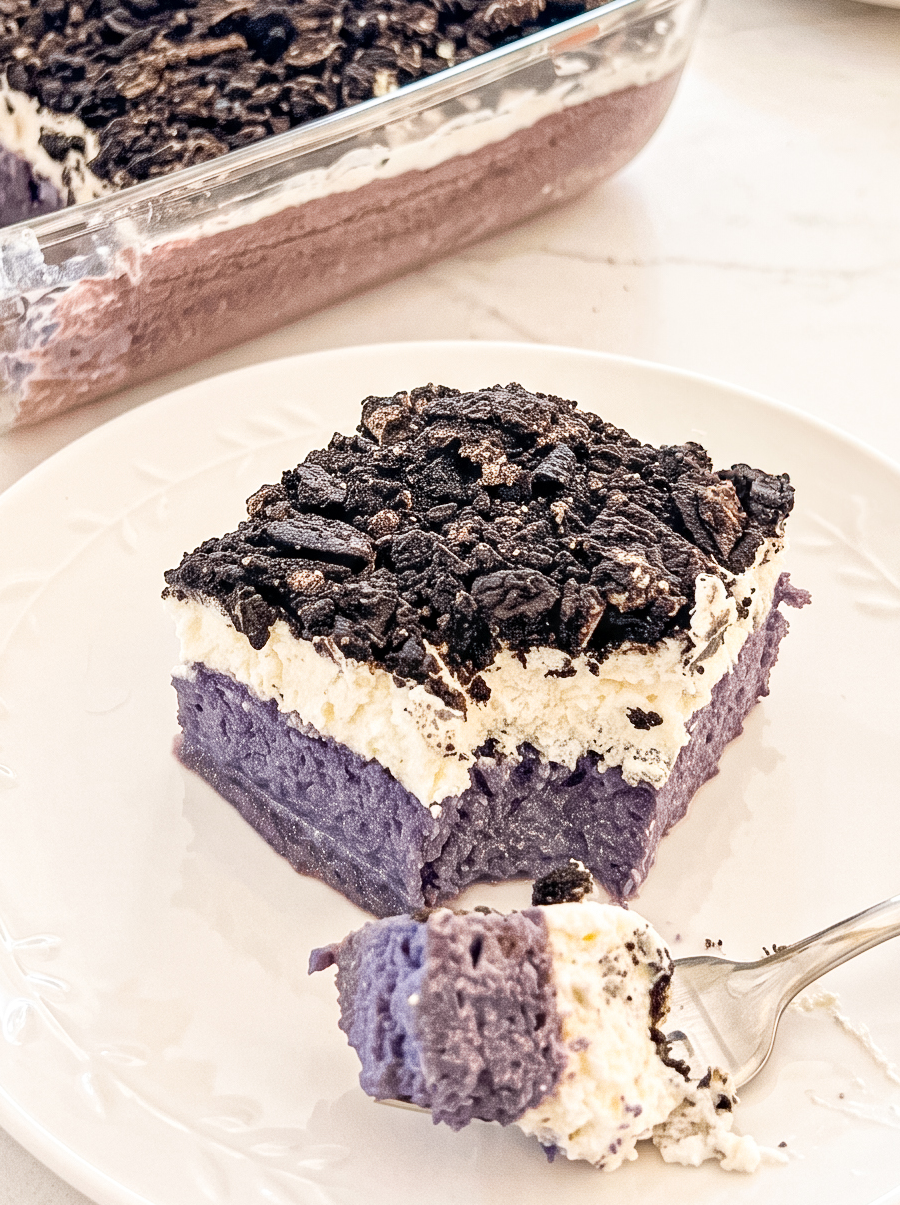 A square slice of ube tres leches on a white plate, with purple cake, white cream, and crushed cookies on top. A fork has removed a bite.
