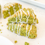 Triangular green matcha scones with white icing drizzle are scattered on a white counter, with a stack in the foreground and more on a tray.