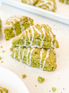 Triangular green matcha scones with white icing drizzle are scattered on a white counter, with a stack in the foreground and more on a tray.