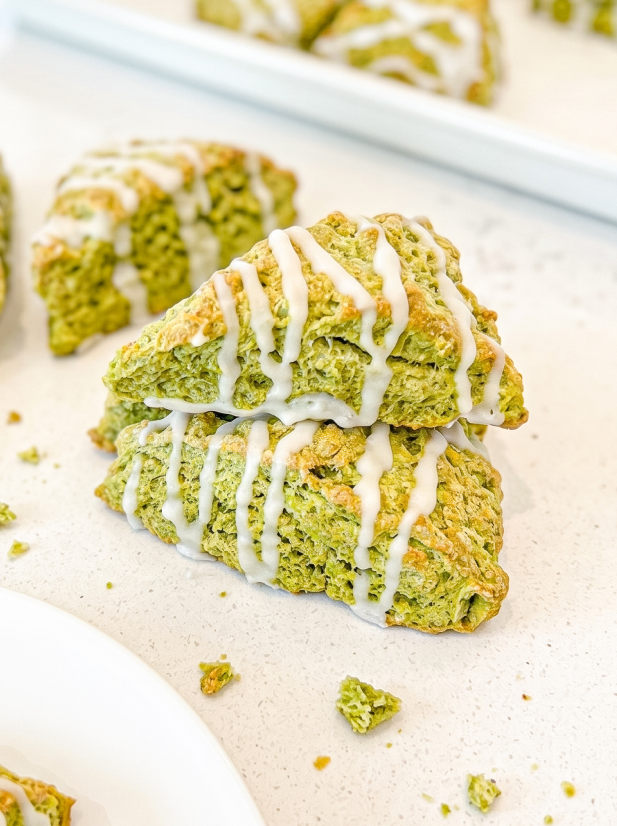 Triangular green matcha scones with white icing drizzle are scattered on a white counter, with a stack in the foreground and more on a tray.