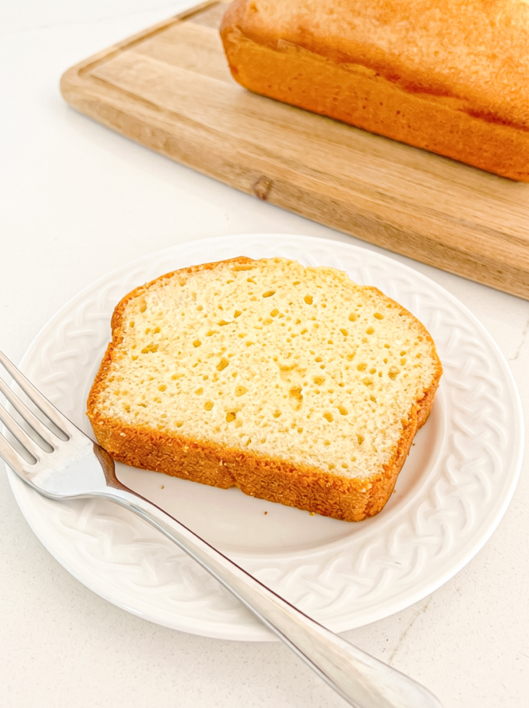 A slice of pound cake rests on a white plate next to a fork, with a whole loaf visible behind it on a wooden cutting board.