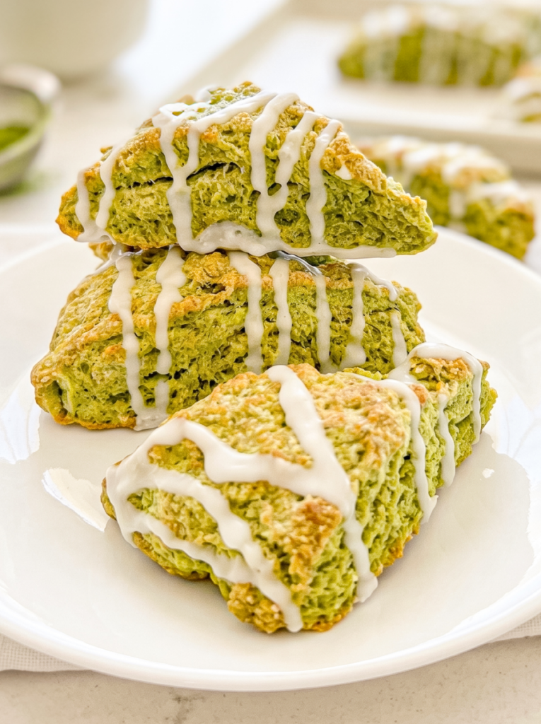 A stack of three triangular matcha scones, drizzled with white icing, sits on a white plate with more scones in the background.
