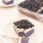 A slice of ube tres leches with a purple cake base, white cream, and crushed dark cookies on a white plate, with a fork taking a piece.