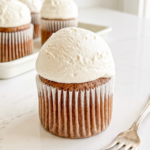 A single Hokkaido mocha chiffon cupcake with a white paper liner and a large dome of white frosting sits on a white marble surface next to a fork, with more cupcakes in the background.