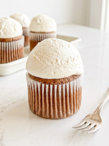 A single Hokkaido mocha chiffon cupcake with a white paper liner and a large dome of white frosting sits on a white marble surface next to a fork, with more cupcakes in the background.