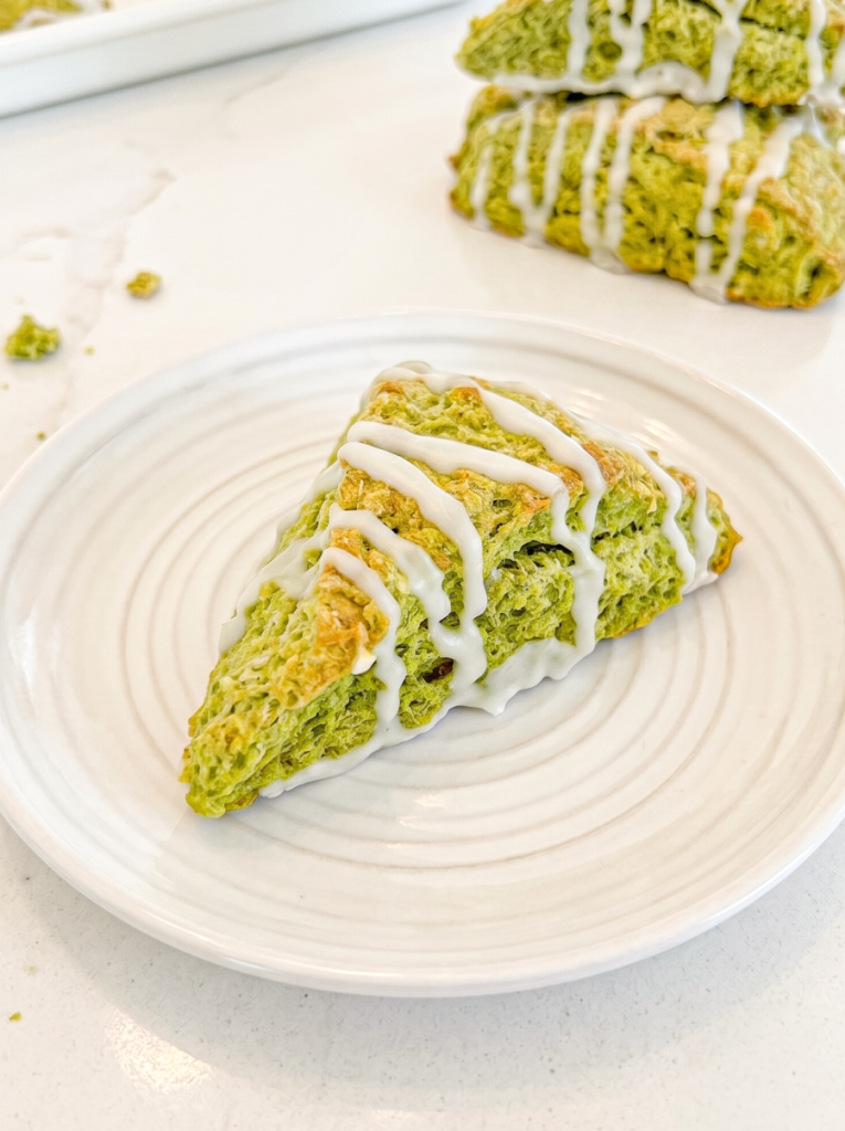 A triangular matcha scone with white glaze on a white plate, with more scones visible in the background.