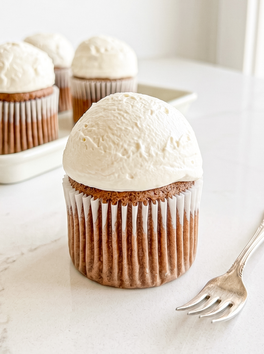 A single Hokkaido mocha chiffon cupcake with a white paper liner and a large dome of white frosting sits on a white marble surface next to a fork, with more cupcakes in the background.