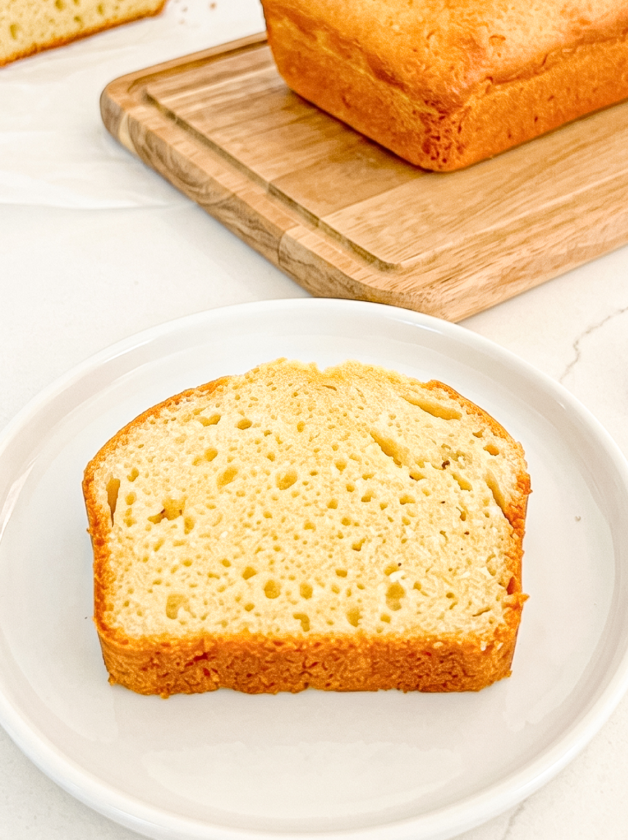A slice of pound cake with a golden-brown crust and pale yellow interior rests on a white plate, with a full loaf on a wooden board behind it.