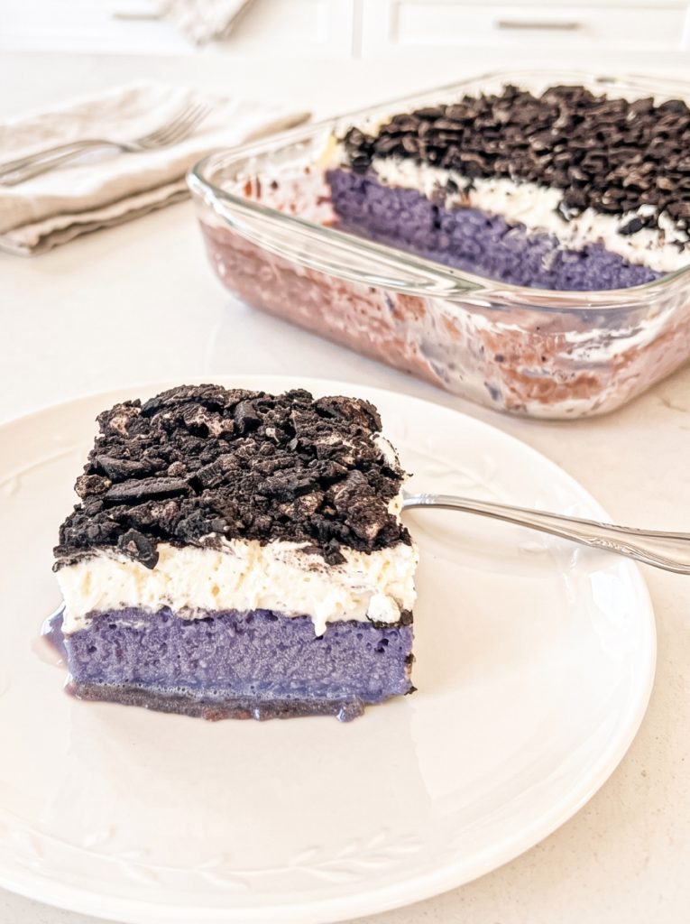 A slice of ube tres leches cake with purple cake, white cream, and crushed dark cookies sits on a white plate, with the rest of the cake in a glass dish behind it
