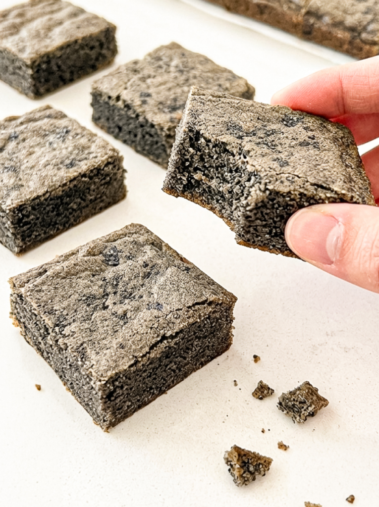 Hand holding a square black sesame blondie, showing its dark, speckled crumb, with several other blondies on a white surface.