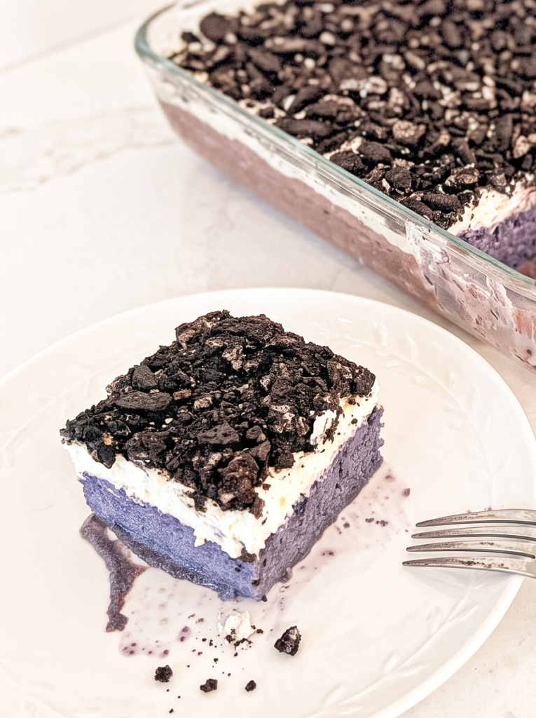 A slice of purple ube tres leches cake with a white topping and crushed dark cookies sits on a white plate, with the baking dish in the background.
