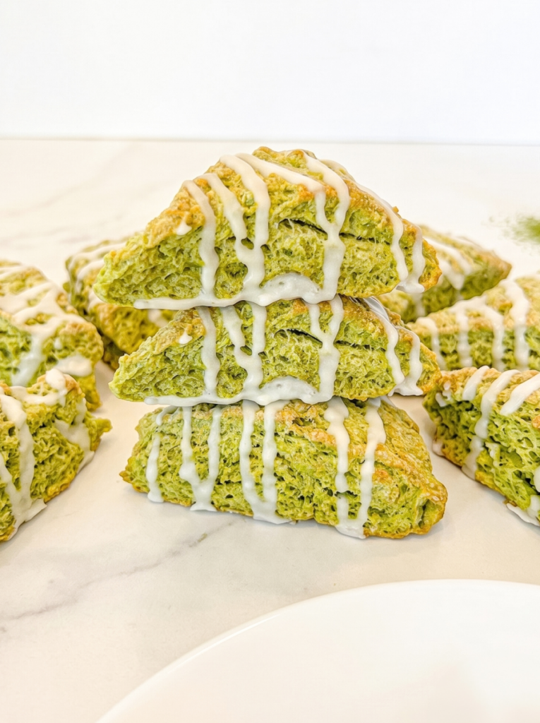 My green matcha scones are stacked and surrounded by more scones, all drizzled with white icing on a marble surface.