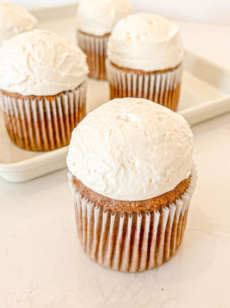 A close-up shows a mocha chiffon cupcake with white frosting, and several others are out of focus on a white tray behind it.