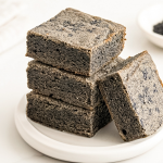 Four square black sesame blondies are stacked on a white plate, with one leaning against the stack. Black sesame seeds are in a small bowl behind them.