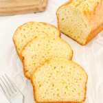A sliced pound cake loaf with a golden-brown crust and pale yellow interior rests on white parchment paper next to three slices and a fork.