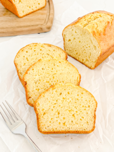 A sliced pound cake loaf with a golden-brown crust and pale yellow interior rests on white parchment paper next to three slices and a fork.