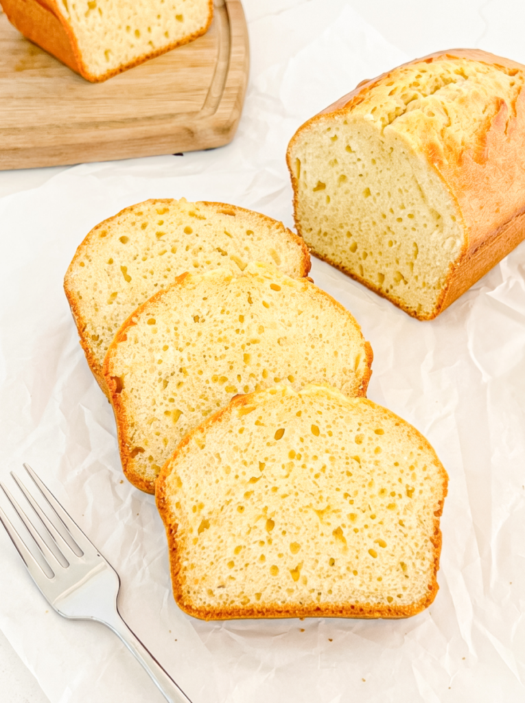 A sliced pound cake loaf with a golden-brown crust and pale yellow interior rests on white parchment paper next to three slices and a fork.
