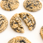 Several round black sesame peanut butter cookies are on a white surface, with one cookie broken in half to show its interior.