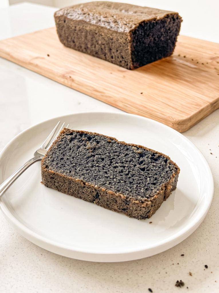 A slice of dark grey black sesame pound cake sits on a white plate with a fork, with a whole loaf on a wooden board in the background.