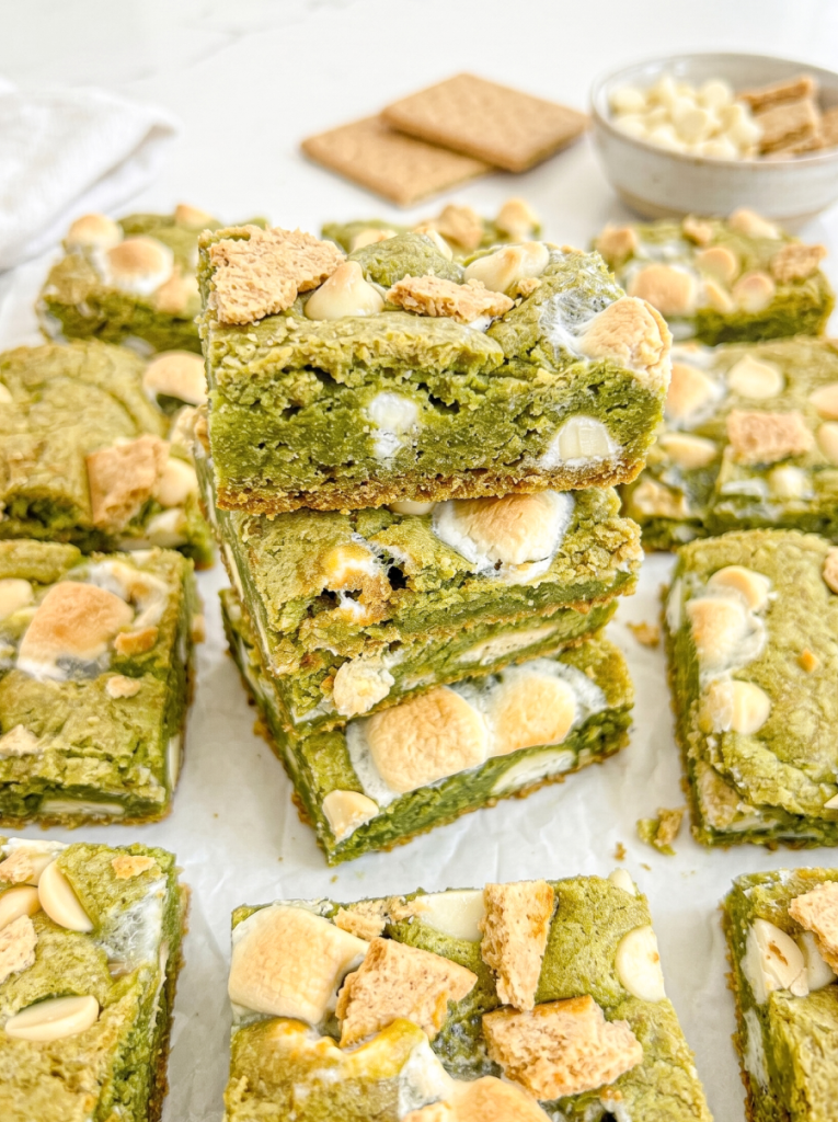 A stack of green matcha s'mores bars with white chocolate chips, toasted marshmallows, and graham cracker pieces, surrounded by more bars.