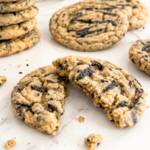 A black sesame peanut butter cookie, broken in half, rests on a white marble surface with a stack of whole cookies behind it.
