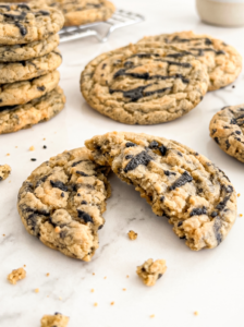 A black sesame peanut butter cookie, broken in half, rests on a white marble surface with a stack of whole cookies behind it.