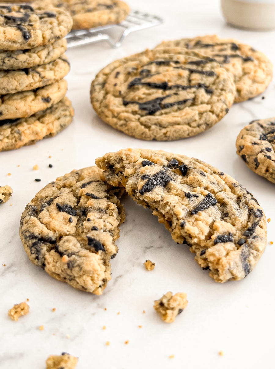 A black sesame peanut butter cookie, broken in half, rests on a white marble surface with a stack of whole cookies behind it.