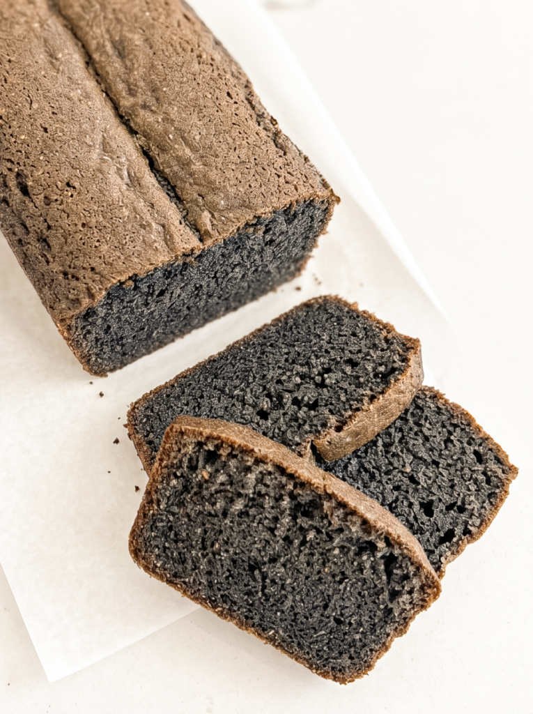 A black sesame pound cake loaf with a brown top crust and dark grey interior sits on white parchment paper next to three cut slices.