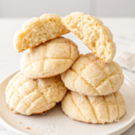 A stack of five sugar-dusted melon pan cookies sits on a white plate, one cookie broken open to reveal its airy interior.