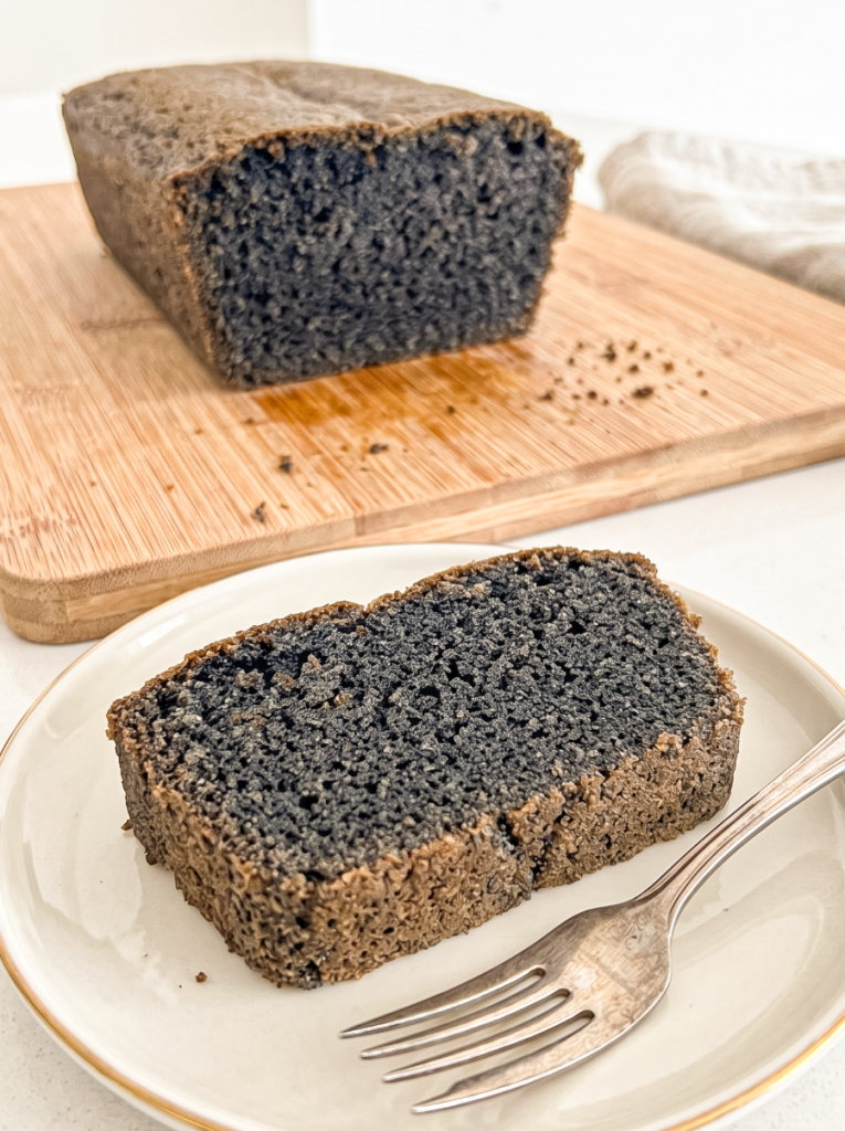 A slice of black sesame pound cake on a plate with a fork, and the loaf on a cutting board behind it.