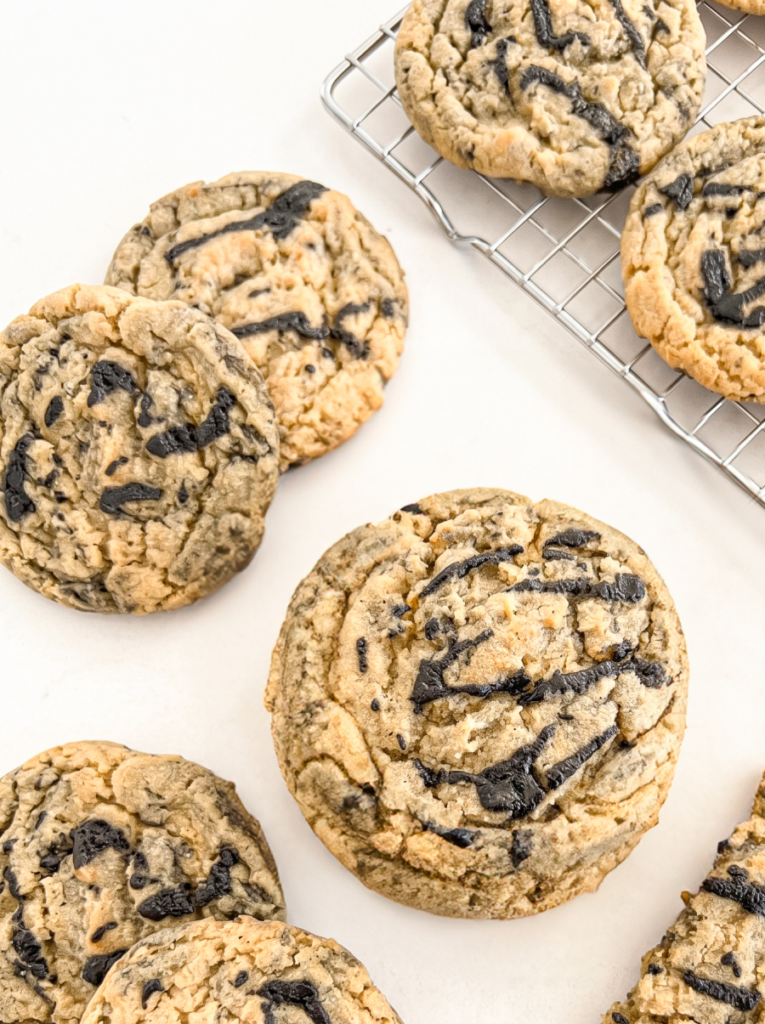 Several round, light brown cookies with dark black sesame drizzles rest on a white surface and a wire cooling rack.