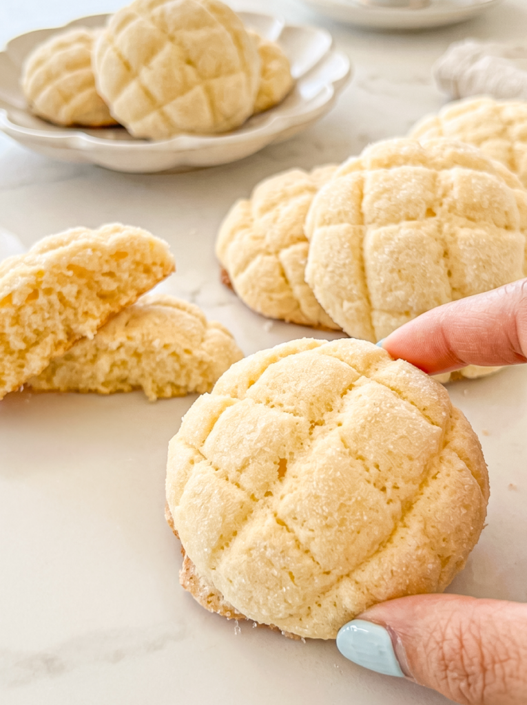 Fingers holding a round melon pan cookie with a criss-cross pattern, surrounded by more cookies on a white marble surface and a plate.