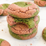 A stack of three cookies, marbled with green matcha and pink strawberry dough, sits on a white marble surface; one has a bite taken out.