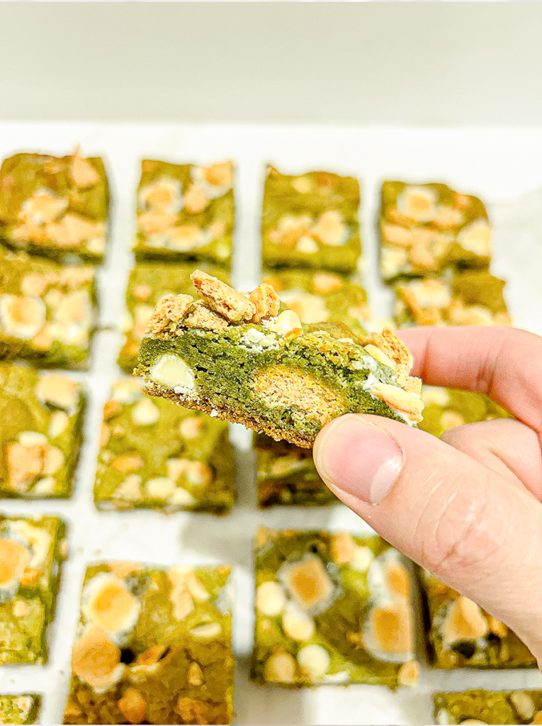 I hold up a square matcha bar with white chocolate chips, melted marshmallow, and graham cracker pieces, with more bars in the background.