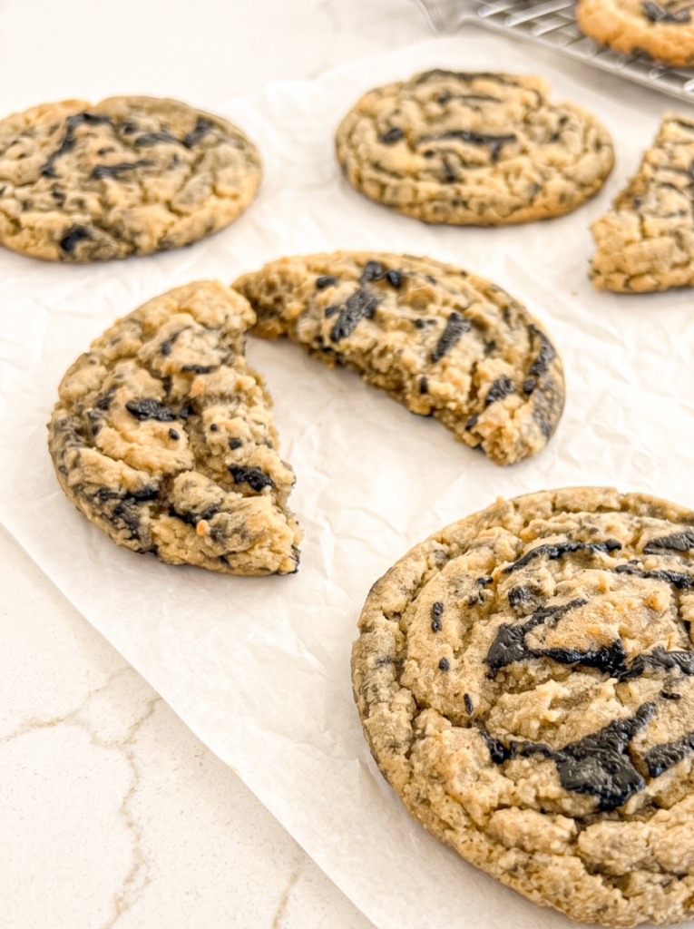 Several round, light brown black sesame peanut butter cookies are on white parchment paper, with one cookie broken in half in the foreground.