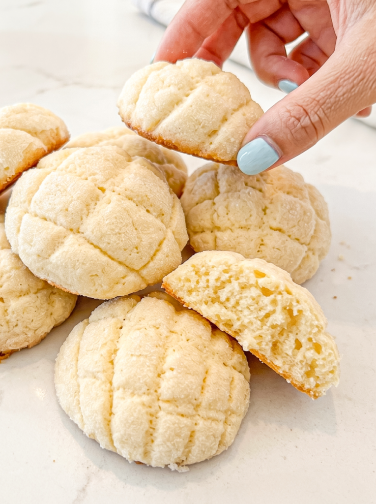 Hand holding melon pan cookie over a pile of cookies with a criss-cross pattern; one cookie is broken to show its soft centre.