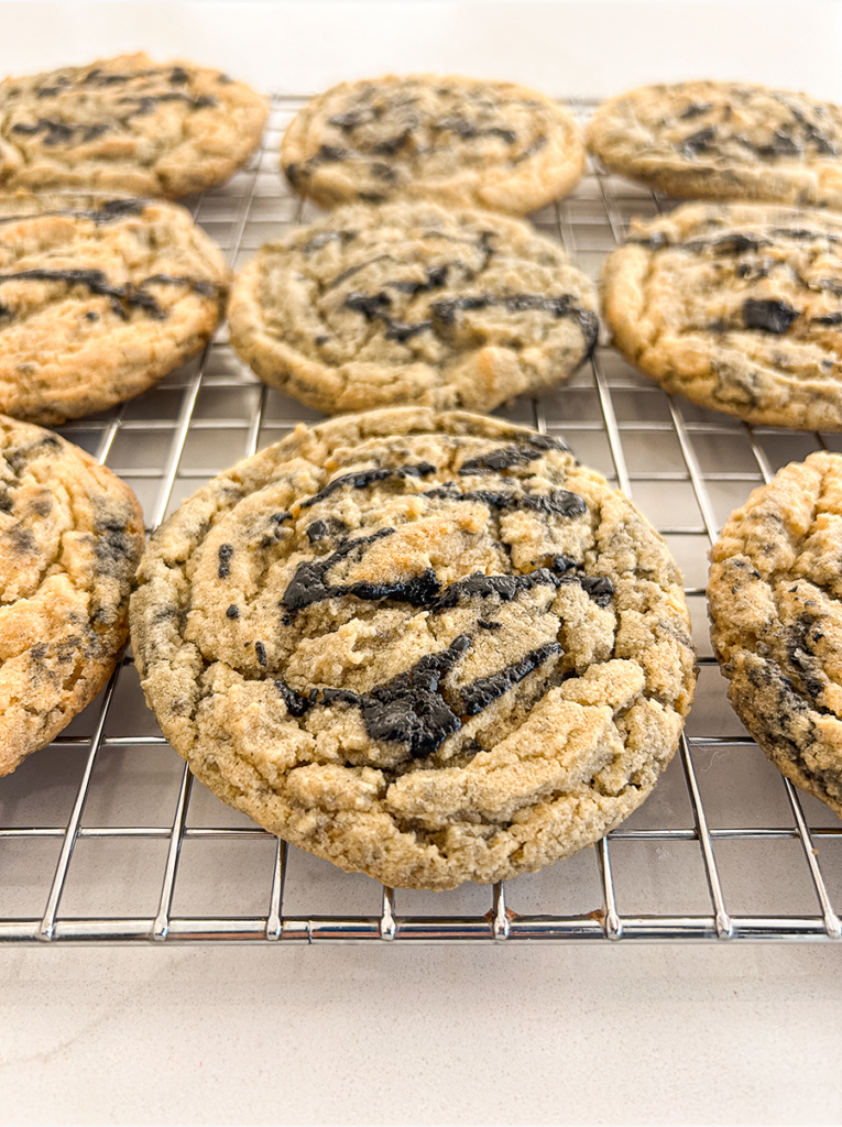 Several round, light brown cookies with dark black sesame streaks and specks sit on a silver wire cooling rack.