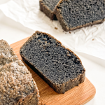 Slices of black sesame pound cake with a dark grey speckled interior are arranged on a wooden board and white parchment paper.