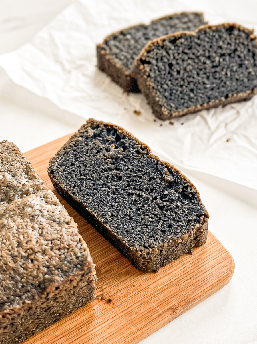 Slices of black sesame pound cake with a dark grey speckled interior are arranged on a wooden board and white parchment paper.