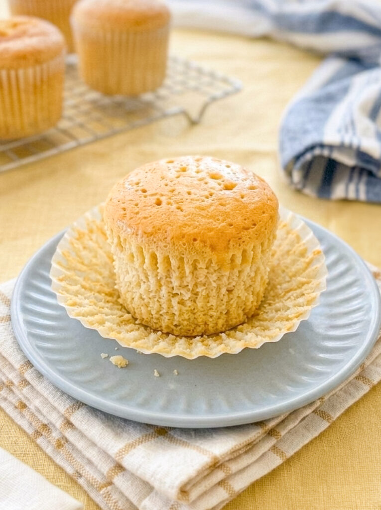A golden honey castella muffin sits on a light blue plate with its paper liner peeled open, revealing a spongy texture.