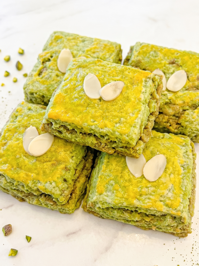 Square pistachio matcha scones are topped with a yellow glaze and almond slivers, arranged on a white surface with scattered pistachios.