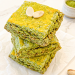 A stack of three square green matcha scones with golden tops, garnished with two almond slivers, rests on white parchment paper.